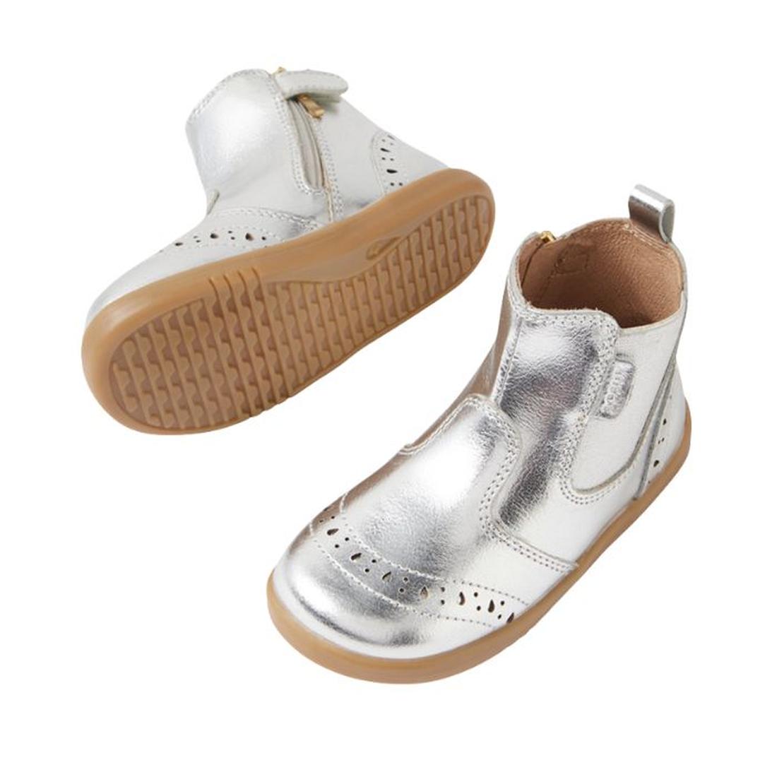The Bobux IW Iwalk Billie Jodhpur Girls Leather Boot in silver features shiny leather, light brown flexible soles, a side zipper, and decorative perforations. One boot stands upright while the other shows its sole., 9420074856302, 9420074856319, 942007485