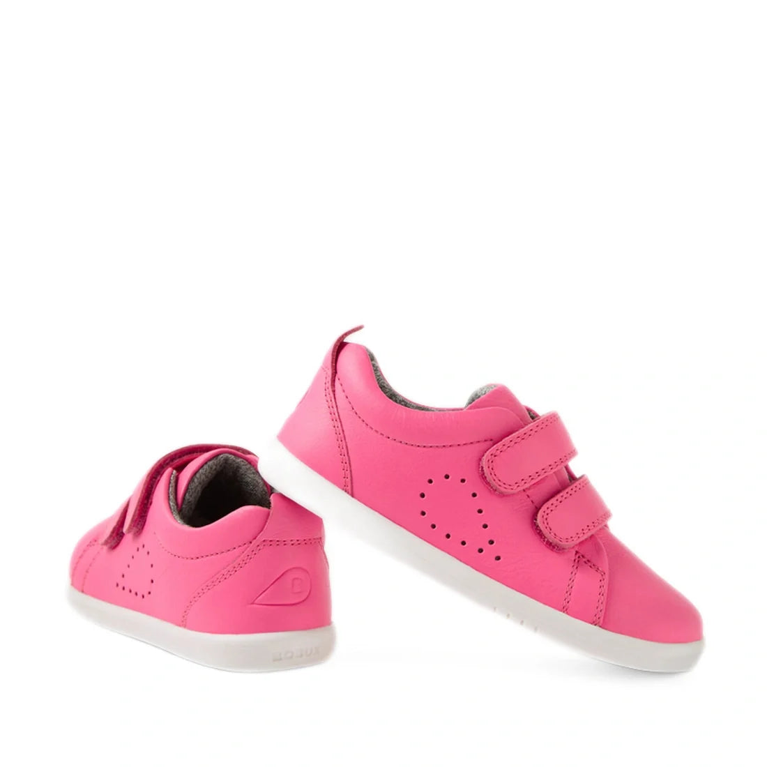 Two vibrant pink Bobux Iwalk Toddlers Grass Court Pink Lemonade Leather Trainers with white soles. One shoe stands upright while the other lies on its side. Both feature two velcro straps and delicate perforated patterns on the sides, highlighting the pla