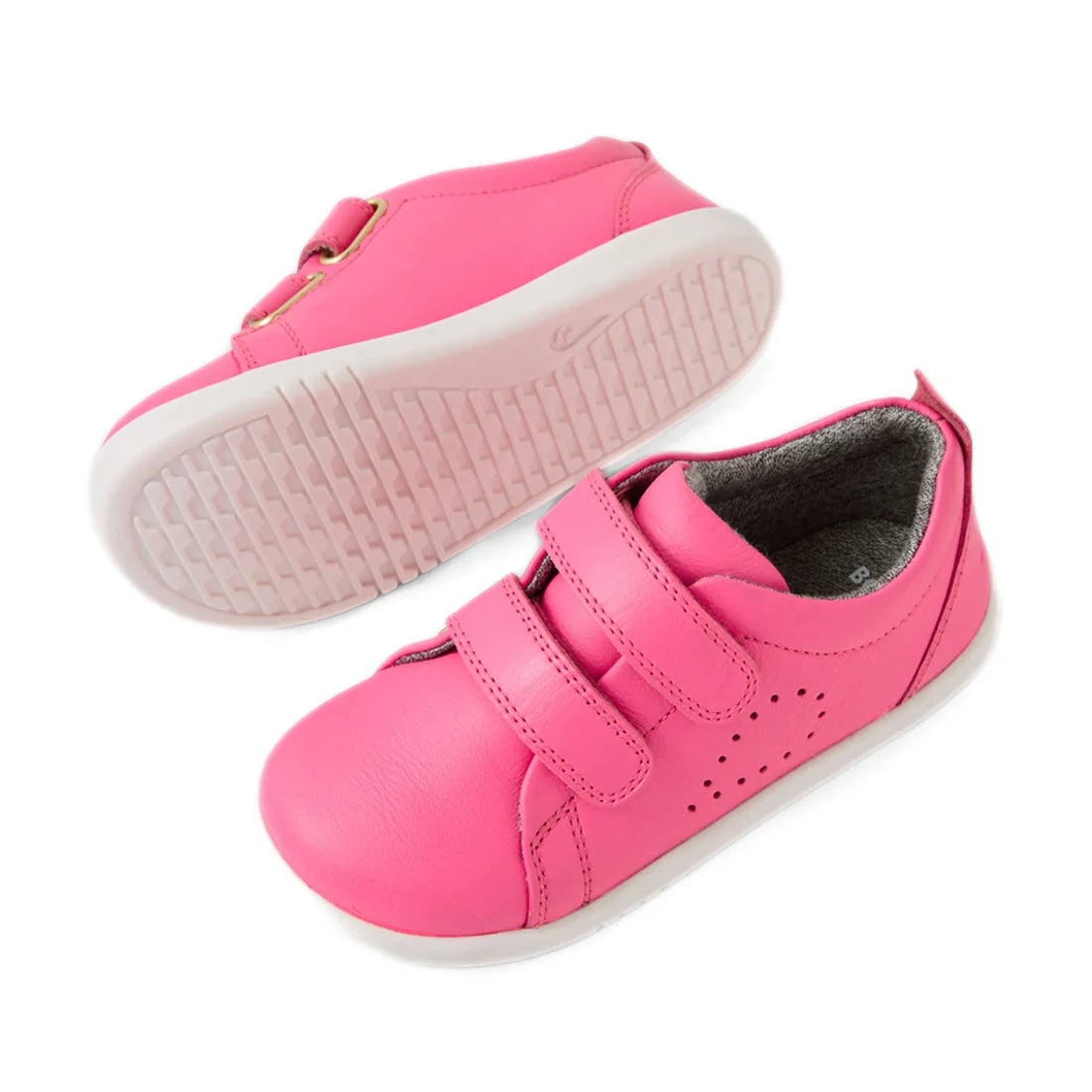 Two vibrant pink Bobux Iwalk Toddlers Grass Court Pink Lemonade Leather Trainers with white soles, one standing upright and the other positioned to display the bottom tread pattern. These kids’ leather sneakers feature two Velcro straps and perforated sid