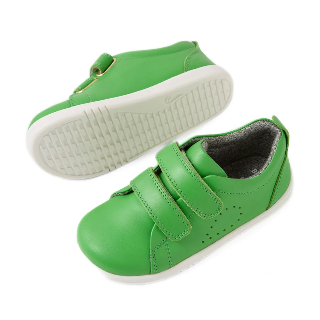 A pair of Bobux Iwalk Toddlers Grass Court Retro Green Leather Trainers, featuring white soles, Velcro straps, and small ventilation holes on the side. One shoe is lying flat while the other is on its side showcasing the sole—perfect toddler trainers for 