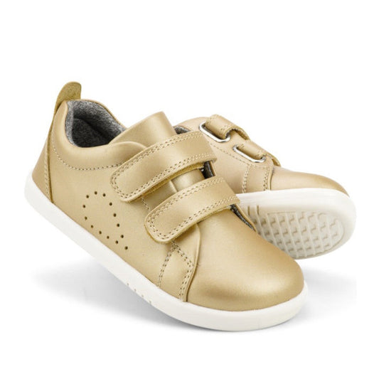 The Bobux Iwalk Grass Court Gold Leather Trainer for kids features shiny gold unisex leather, white soles, and a double strap closure. Displayed on a plain white background, one shoe stands upright while the other is angled behind it.