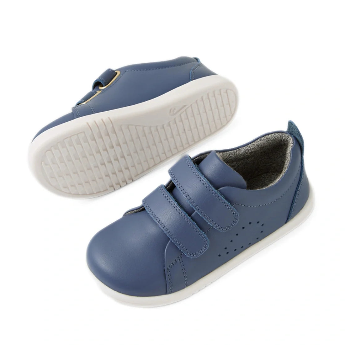 A pair of Bobux Iwalk Toddlers Grass Court Vintage Indigo Blue Leather Trainers is displayed—one toddler sneaker stands upright while the other rests on its side. The shoes feature white soles, Velcro straps, side perforation details, and a soft gray inne