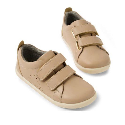 The Bobux IW Iwalk Grass Court Leather Sneaker in Warm Taupe is a beige toddler boy's shoe with a premium leather upper, white soles, and two Velcro straps per shoe, displayed on a white background.