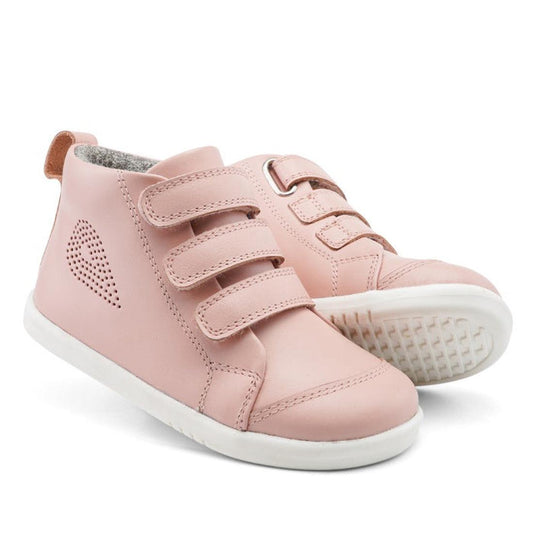 The Bobux IWalk IW Hi Court Boot in Seashell Pink is a toddler shoe with white soles, premium coated leather, three hook-and-loop straps, and a small heart-shaped cutout on the side.