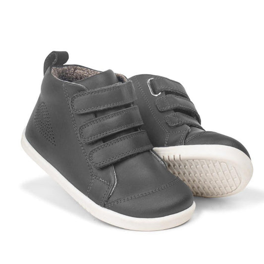 The Bobux IWalk IW Hi Court Boot in Smoke Grey, featuring three Velcro straps and white rubber soles, is displayed against a white background—one boot upright and the other tilted to reveal its sole.