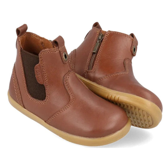 A pair of Bobux IWalk Jodhpur Boot Toffee 620826A kids leather boots, featuring side zippers, elastic panels, and tan rubber soles. One boot is upright while the other leans to reveal the textured sole. Free shipping NZ wide.