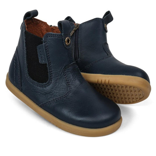 The Bobux Iwalk Jodhpur Ankle Boots Navy 620825A feature black elastic side panels and tan rubber soles—perfect stylish leather boots for toddlers, with one boot upright and the other resting on its side.