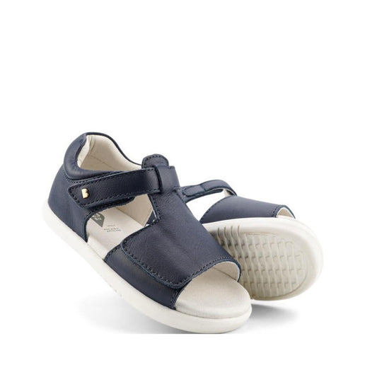 The Bobux IW Iwalk Mirror Navy Kids Sandal 633429 features navy leather, open toes, white soles, adjustable Velcro straps, and a small metal side accent. One sandal is propped against the other on a white background.