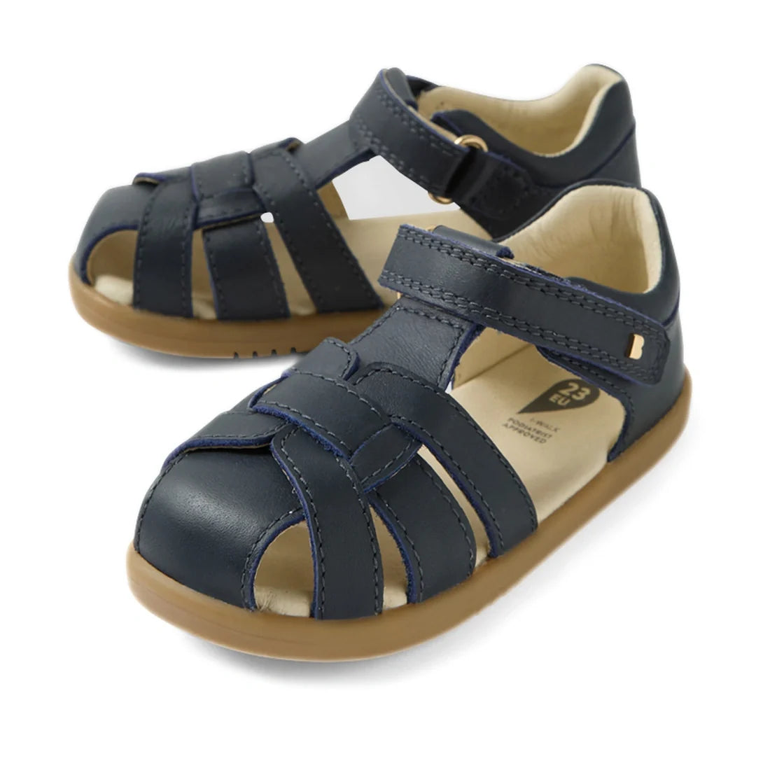 The Bobux IWalk Skip Twist Sandal Navy Closed Toe Toddlers are a pair of navy blue leather sandals for toddlers, featuring a closed toe, Velcro straps, tan soles, and beige lining. Displayed on a white background, these sandals combine style and protectio