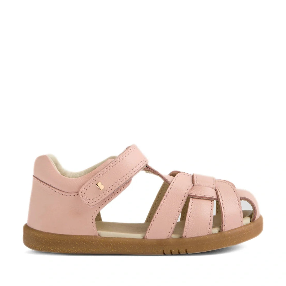 The Bobux IWalk Skip Twist Sandal in Cameo Rose Pink features a closed heel, open toe, woven leather straps, an adjustable Velcro ankle strap, and a brown rubber sole. Shown from the side against a white background, this charming sandal is an excellent ch