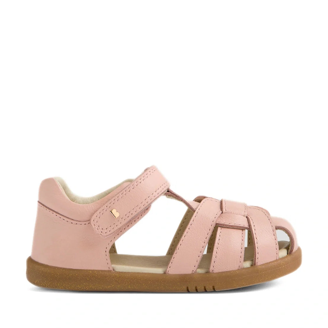 The Bobux IWalk Skip Twist Sandal in Cameo Rose Pink features a closed heel, open toe, woven leather straps, an adjustable Velcro ankle strap, and a brown rubber sole. Shown from the side against a white background, this charming sandal is an excellent ch
