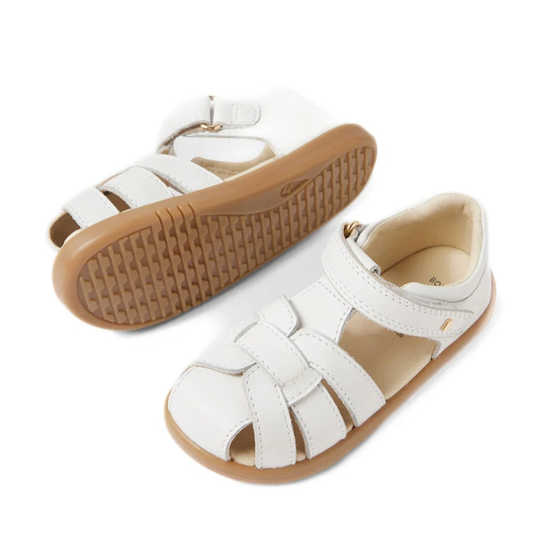 Introducing the Bobux IWalk Skip Twist Sandal in White, a pair of closed-toe leather sandals for toddlers featuring woven white leather straps over the toes, secure velcro ankle straps, and durable tan rubber soles. These breathable kids’ sandals are desi