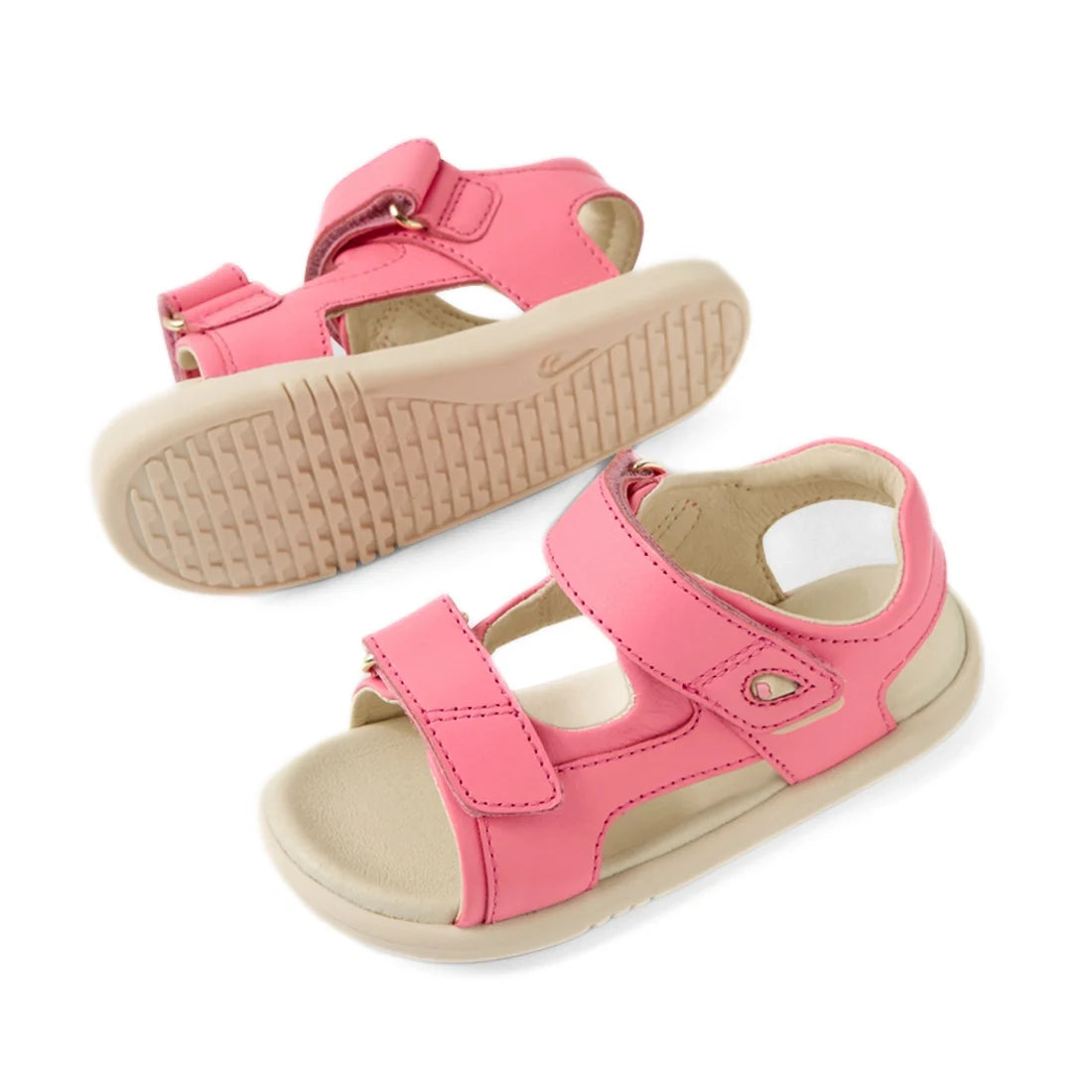 A pair of Bobux IWalk Strand Sandal Pink Lemonade Open Toe Toddlers, featuring soft pink leather, adjustable Velcro straps, and beige soles, is displayed from the side and bottom against a white background. These washable sandals are crafted for comfort a