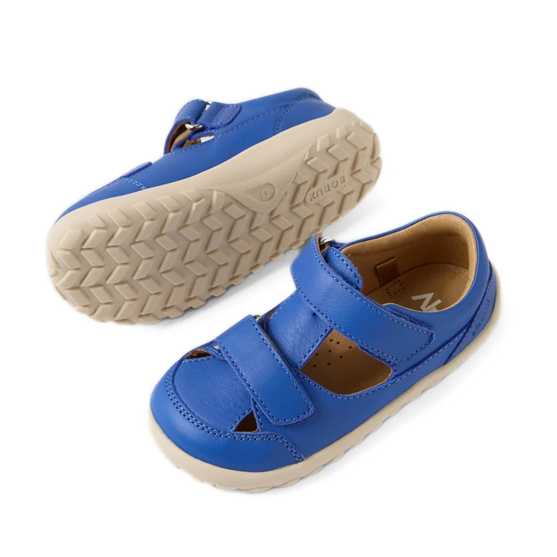 A pair of Bobux IW Iwalk Troop Toddlers Leather Sandal Dazzling Blue Barefoot shoes for children, featuring beige rubber soles and Velcro straps. One shoe stands upright while the other lies on its side, displaying the tread pattern on the sole., 94200748