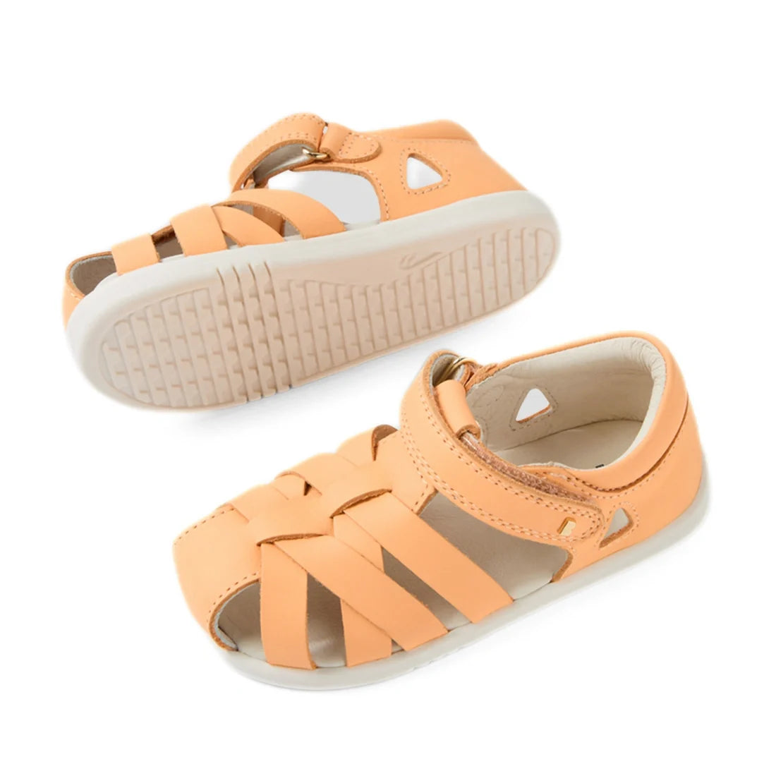 A pair of Bobux Iwalk IW Tropicana II Peach Jelly Leather Sandals, crafted from peach-colored leather with woven straps, ankle buckles, and durable white rubber soles that provide closed-toe protection. One sandal is displayed sole-up while the other rest