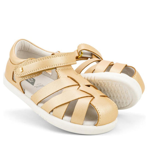 A pair of Bobux Iwalk IW Tropicana II Sandal 638311 in Pale Gold for toddlers, featuring closed toes, woven Quick Dry Leather straps, and white rubber soles. One sandal stands upright; the other leans to reveal its textured sole.