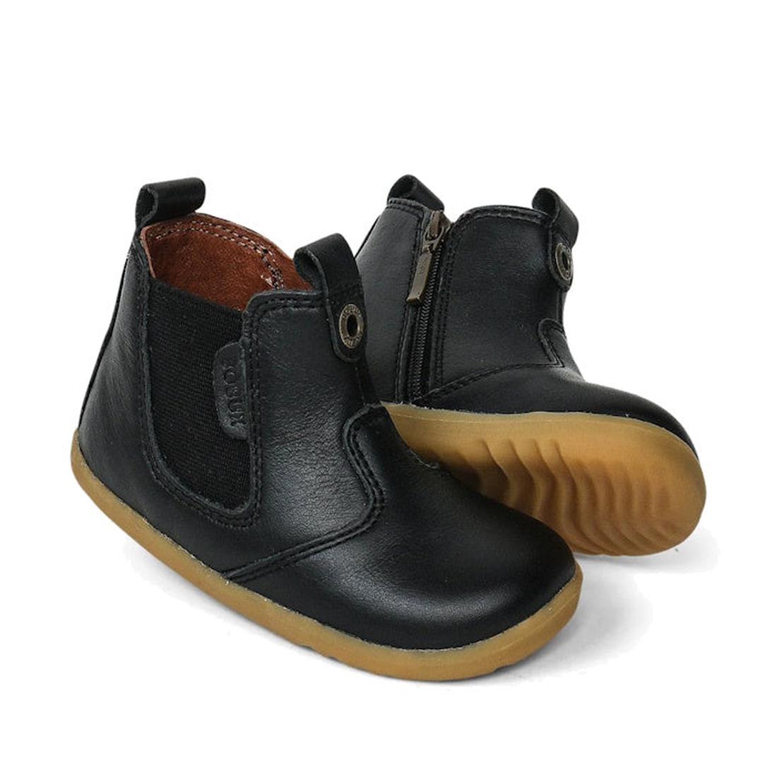 The Bobux Step Up Jodphur Boot Black 721928A is a unisex toddler boot in black leather, featuring elastic side panels, side zippers, and light brown textured rubber soles. One boot stands upright, the other leans to display its sole.