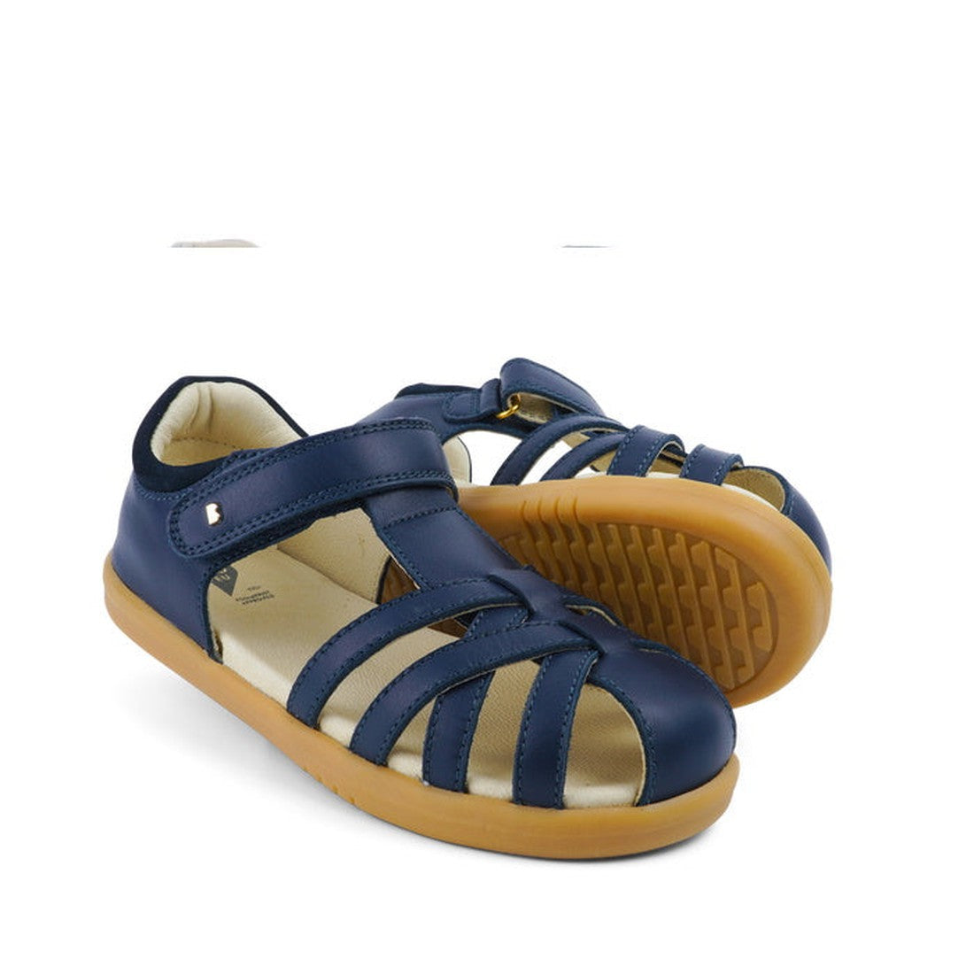 The Bobux Kids + Plus Girls Cross Jump Navy Leather Sandal 837003 features closed toes, open sides, tan rubber soles, and an adjustable strap. One sandal stands upright while the other rests on its side.