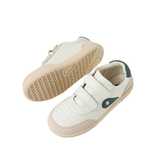 The Bobux Kids Plus Junior KP Champ II Sneaker in Chalk White and Rainforest Green features leather uppers, beige soles, green heel accents, velcro straps, a small heart design on the side, and a durable tread. One shoe is upright; the other shows the sol