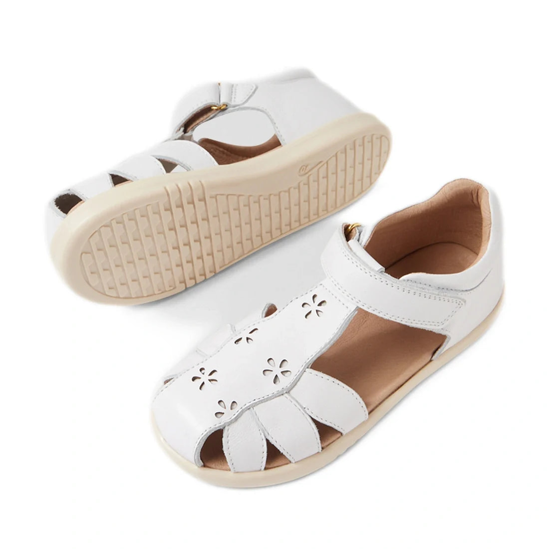 A pair of Bobux Kid+ Plus KP Holly White Closed Toe Sandals for girls featuring white leather, cutout flower designs, beige soles, and Velcro straps. One sandal stands upright while the other is positioned on its side to display the sole’s tread pattern—t