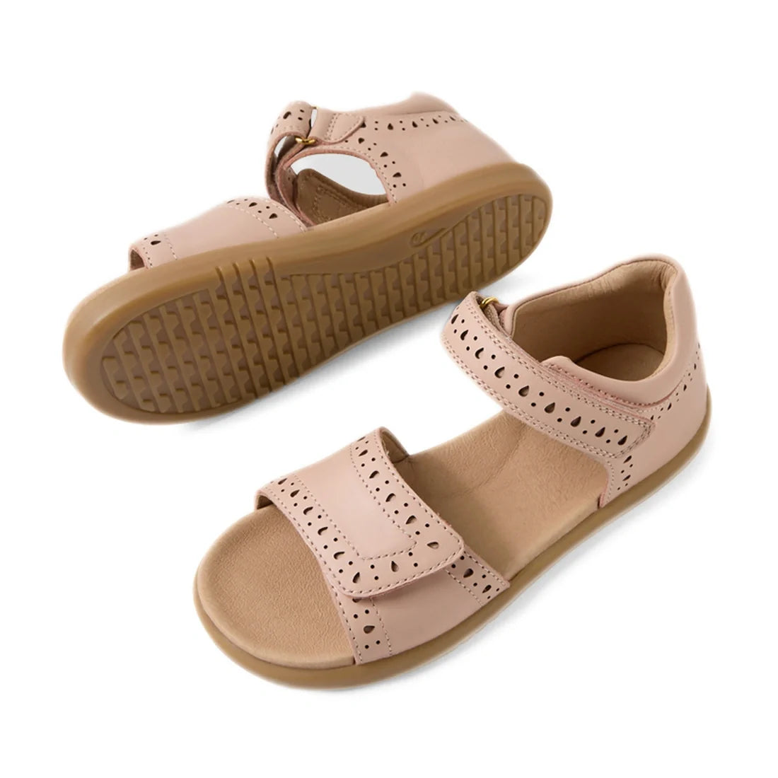 Two Bobux Kid+ Plus KP Logan Cameo Rose Pink Open Toe Sandals are displayed—one upright and one lying on its side to showcase the tan sole. Featuring decorative perforations, adjustable Velcro straps, and crafted from stylish pink leather, these sandals b