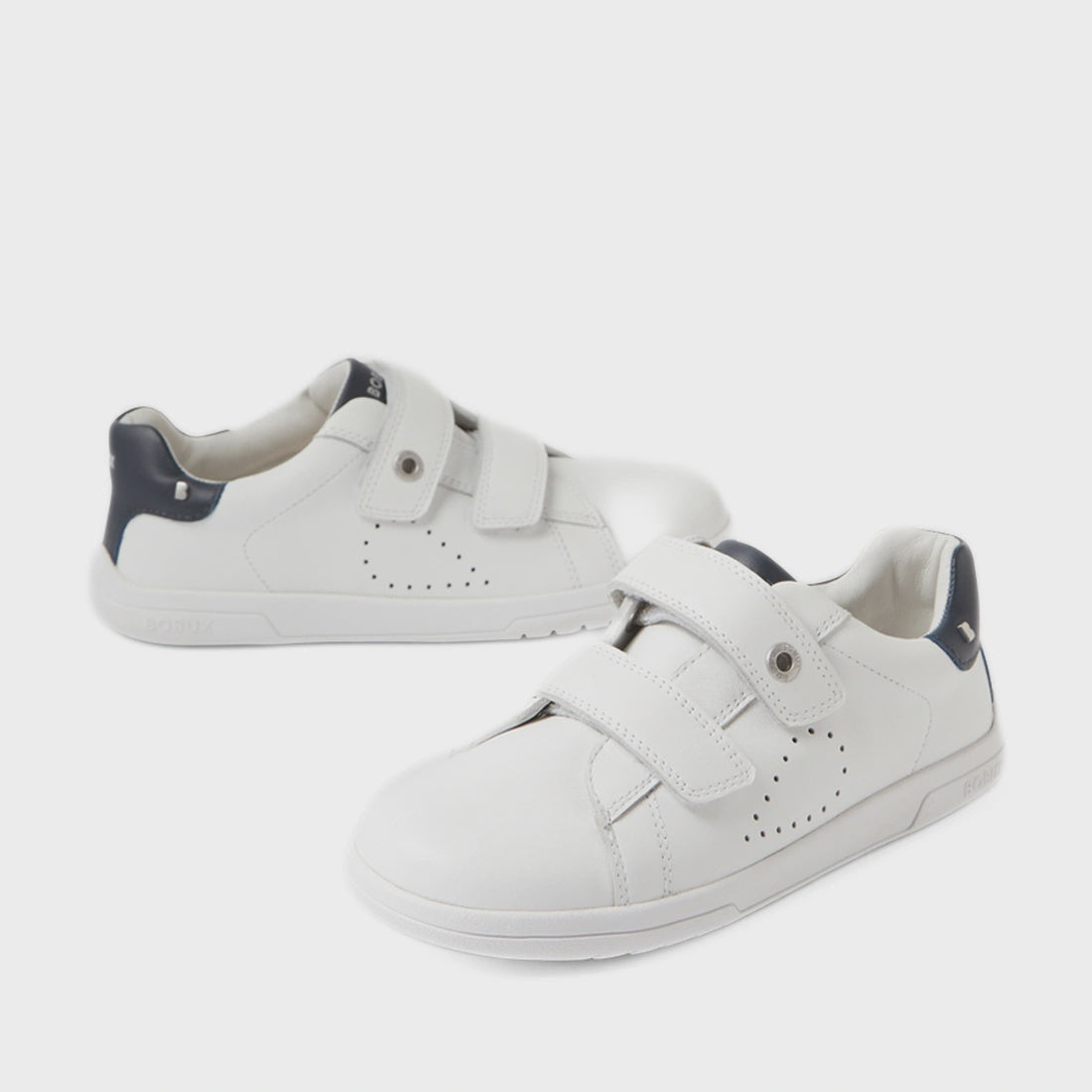 A pair of Bobux Kid+ Plus KP Maverick Leather Sneakers in White Navy—children’s sneakers with adjustable velcro straps, dark blue heel accents, and crafted from breathable leather for comfort—are stylishly displayed on a white background.