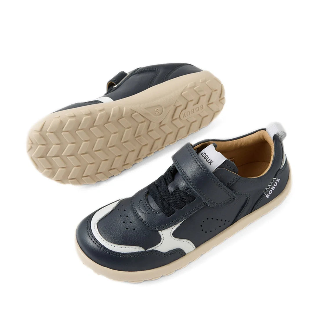 A pair of Bobux Kid+ Plus KP Neo Leather Sneaker Navy Alpha for kids is shown from the side and bottom. These navy blue leather sneakers feature white soles and accents, a barefoot-shaped toebox, a Velcro strap, and toe perforations for enhanced breathabi