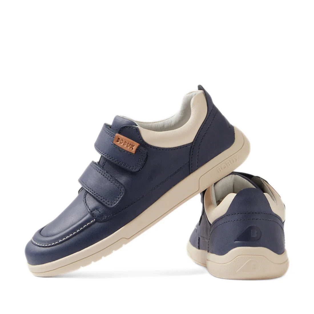 A pair of Bobux Kid+ Plus KP Oakley Leather Sneakers in Vintage Indigo Blue for kids, featuring two velcro straps, a beige and navy blue leather upper, a light-colored sole, and a brown side label reading BOFINX. With styling reminiscent of a classic boat