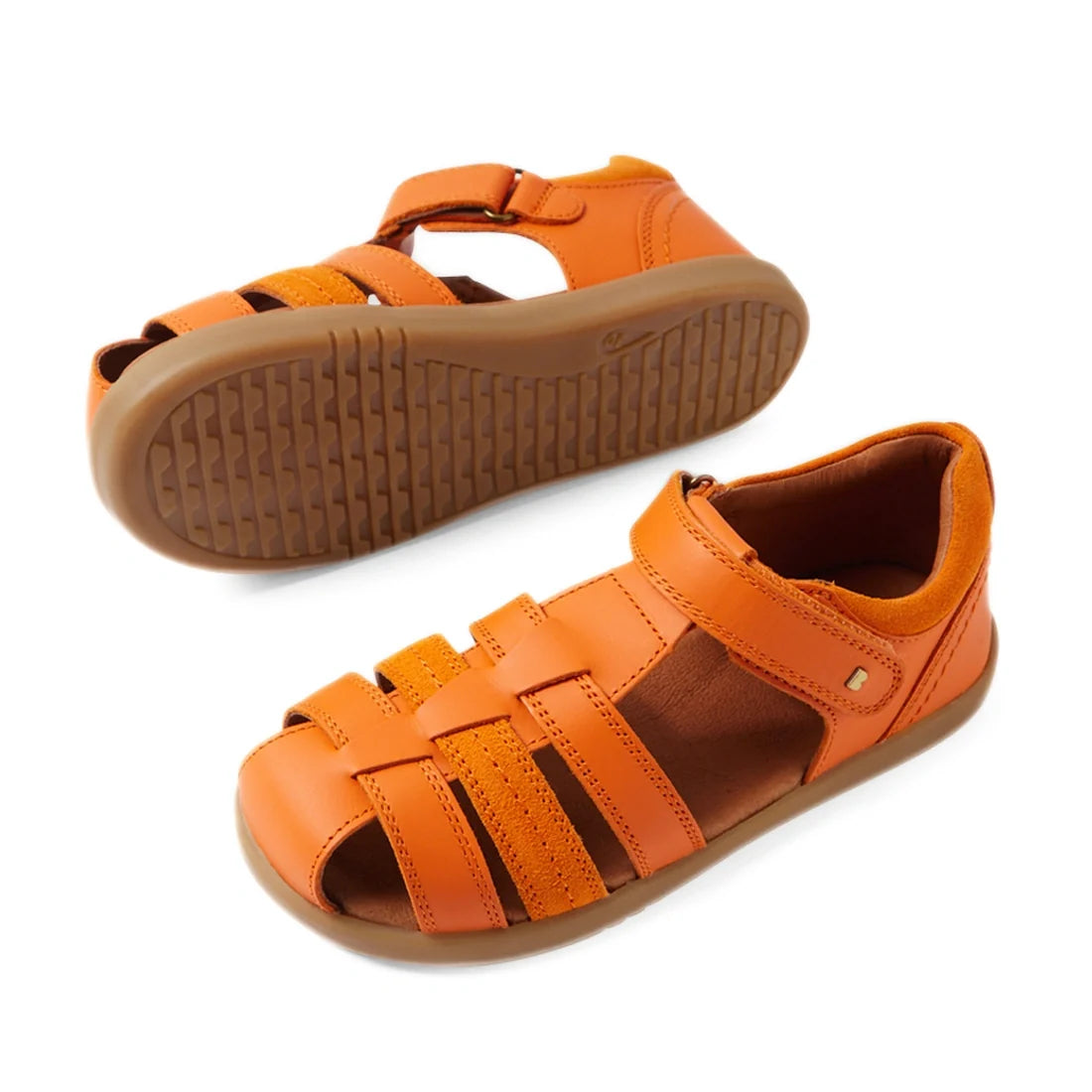Introducing the Bobux Kid+ Plus Roam Orange Peel Closed Toe Sandal: vibrant orange leather sandals perfect for kids’ summer adventures. These stylish shoes feature secure velcro straps and durable brown rubber soles. One sandal stands upright while the ot