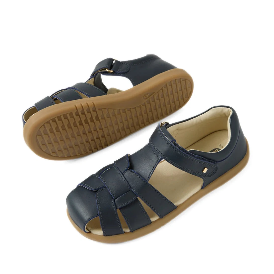 A pair of Bobux Kids Plus KP Skip Twist Navy Closed Toe Sandals, featuring navy blue uppers and tan rubber soles—one sandal lying flat and the other on its side to highlight the tread pattern and adjustable Velcro ankle strap—providing breathable comfort 