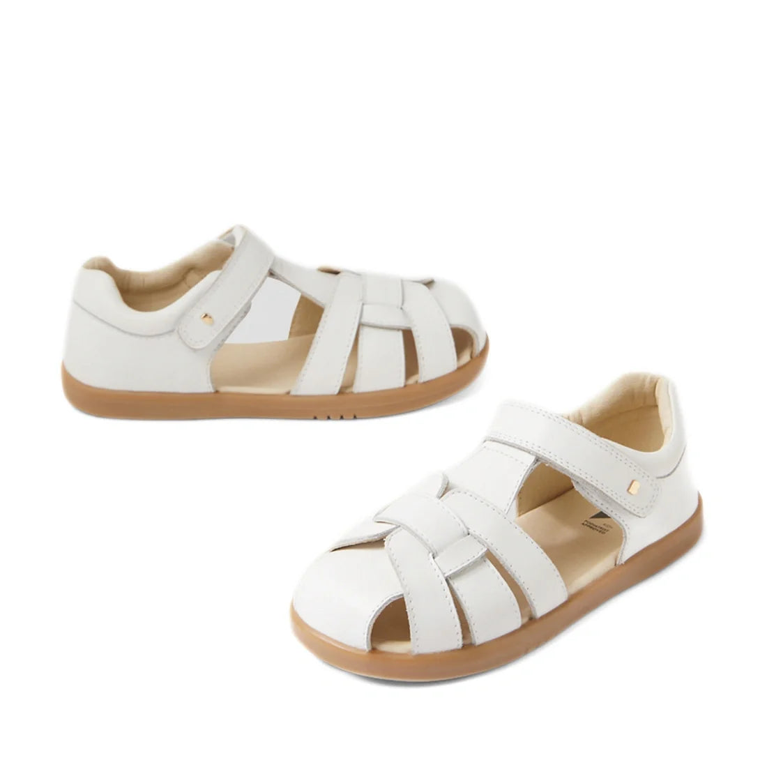 Two Bobux Kids Plus KP Skip Twist White Closed Toe Sandals for children, featuring open sides, tan rubber soles, and adjustable Velcro straps, are displayed on a white background.