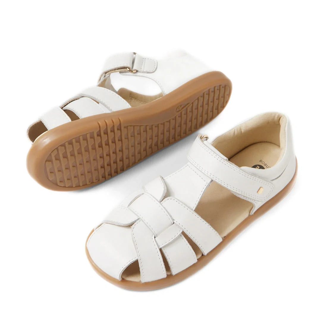 A pair of Bobux Kids Plus KP Skip Twist White Closed Toe Sandals with tan rubber soles, featuring a closed-toe design, multiple straps, and a hook-and-loop ankle strap. One leather sandal is upright, while the other is on its side showing the sole., 94200