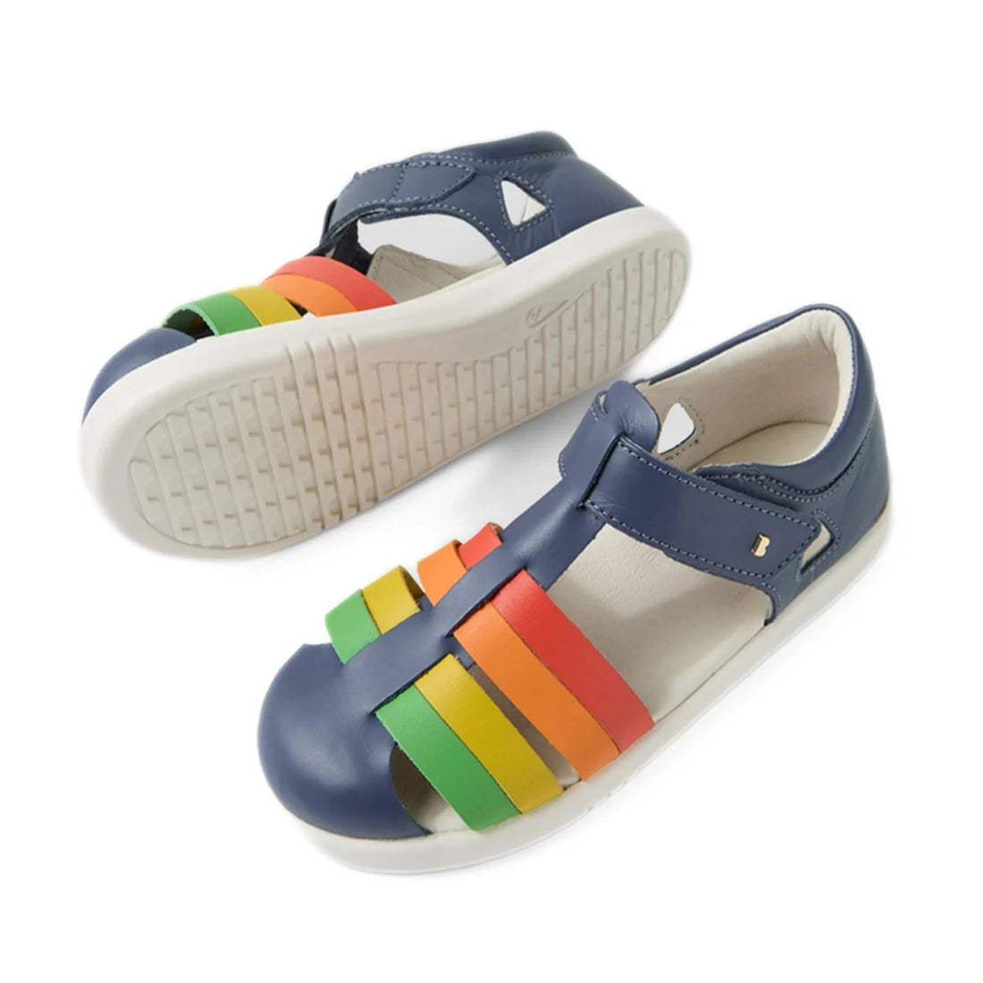 Two pairs of Bobux Kid + Plus Tidal Closed Toe Quick Dry Sandals in Vintage Indigo Blue, designed for kids. These sandals feature blue uppers, water-friendly leather construction, white soles, and colorful straps in green, yellow, orange, and red across t