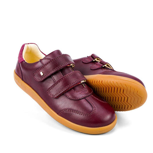The Bobux Kids Plus KP Sprite Sneaker in Boysenberry features a burgundy leather upper, tan rubber sole, unisex style, double hook-and-loop velcro straps, and a low-cut design. One shoe stands upright while the other is on its side.