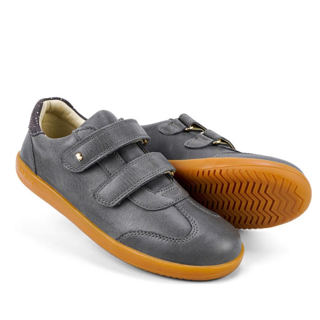 The Bobux Kids Plus KP Sprite Sneaker in Charcoal Grey features two Velcro straps, light brown rubber soles, and a sleek design—perfect unisex shoes for winter 2023, with one shoe upright and the other showing its sole.