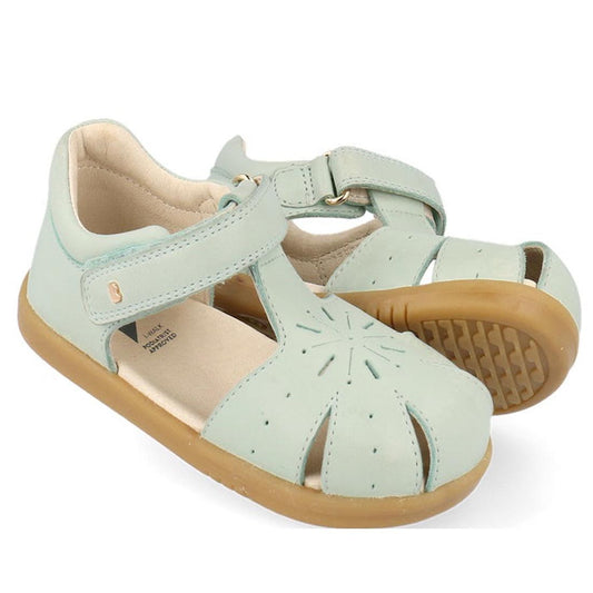 The Bobux Iwalk IW Compass Kids Mist Blue Leather Sandal 641205 features mist blue leather, tan rubber soles, closed toes with cut-out designs, and adjustable hook-and-loop straps. These summer sandals are shown on a white background.
