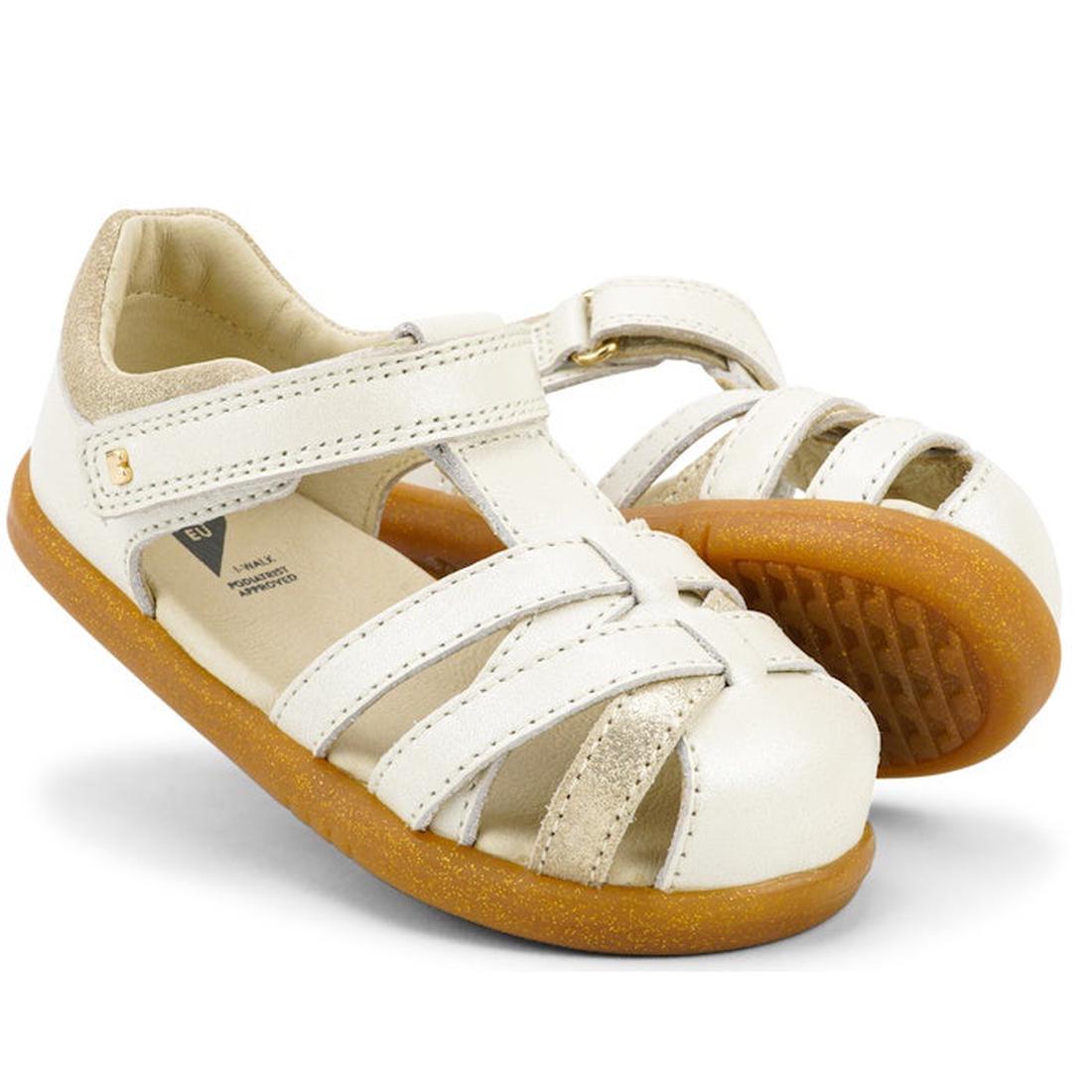 The Bobux iwalk Cross Jump Sandal Moon Pearl Closed Toe 636708 is a kids' sandal with white and gold straps, Velcro fastening, and tan rubber soles. One leather sandal stands upright while the other leans against it.