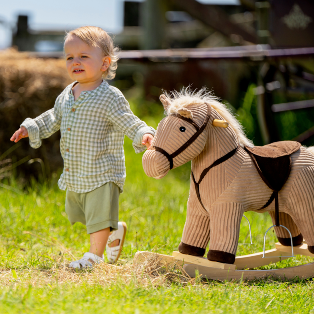 A young child walks outdoors on grass, wearing a light checked shirt, shorts, and Bobux iwalk Cross Jump Sandal White Closed Toe 636706 with Velcro fastening. The child holds the mane of a plush rocking horse under bright sunlight., 9420062083949, 9420062