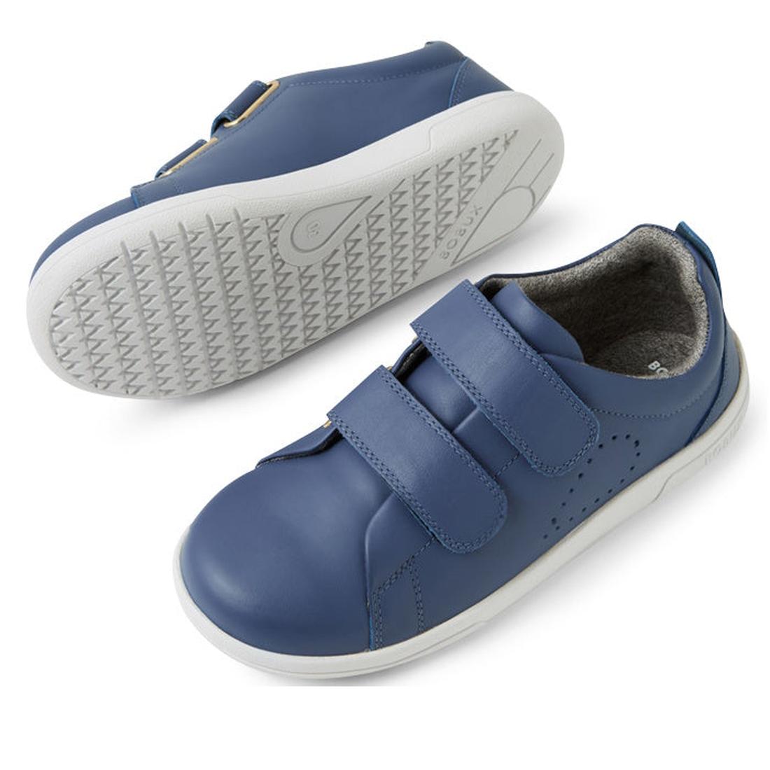 The Bobux KP Kids Plus Grass Court II Leather Sneaker in Vintage Indigo Blue features two adjustable straps, white soles, subtle perforations on the sides, and a textured sole, shown with one shoe upright and the other on its side., 9420074859440, 9420074