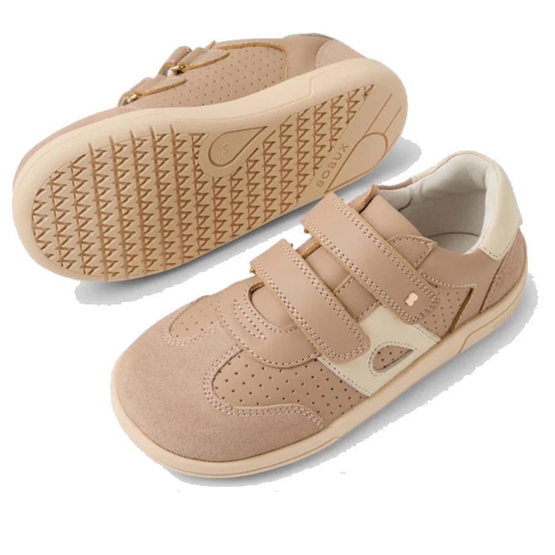 The Bobux KP Kids Plus Titan Juniors Leather Sneaker in Warm Taupe features beige leather, adjustable hook-and-loop straps, and textured slip-resistant soles. One shoe stands upright; the other is flipped to show the sole's tread pattern., 9420074855893, 
