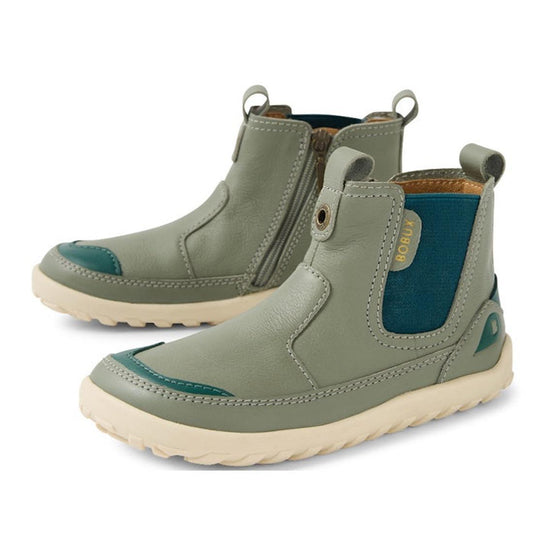 The Bobux KP Kids Plus Trail Leather Boot Juniors in Vetiver Green Alpha features gray-green leather, teal elastic panels, inner ankle zipper, beige soles, barefoot-shaped toe box, pull tabs, and visible BOBUX branding on the side.