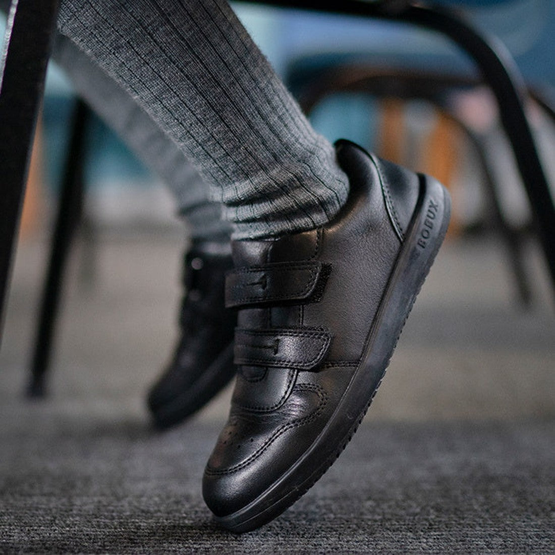 A child in cozy gray socks and Bobux Kid+ LeapTrainer black leather school shoes with sturdy soles and easy fastenings sits on gray carpet.  ksnz, 9420068510494, 9420068510500, 9420068510517, 9420068510524, 9420068510531, 9420068510548, 9420068510555