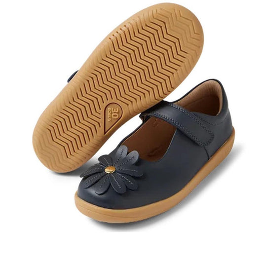The Bobux Kid+ Pixie Girls Mary Jane Leather Navy 850502 features navy leather, tan rubber soles, a Velcro strap, and a large flower motif on the toe; one shoe is upright while the other shows off its textured sole.