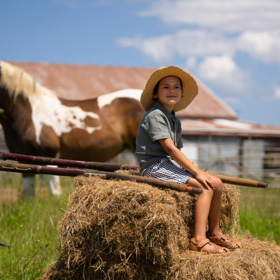 A young child wearing Bobux IWalk Roam Sandal Caramel Closed Toe 626015A and a straw hat sits on a hay bale in a sunny field with a barn and horse in the background, looking content amid peaceful rural farm scenery., 9420062047422, 9420062047439, 94200620