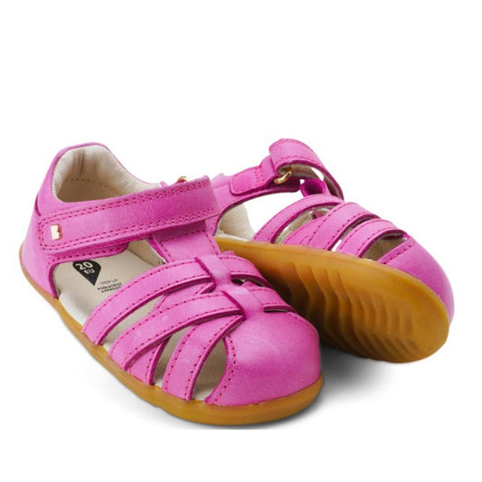 A pair of Bobux Step Up Toddlers Cross Jump Sandal Bright Pink 731109 features closed toes, Velcro straps, and light brown rubber soles. One sandal stands upright while the other leans against it.
