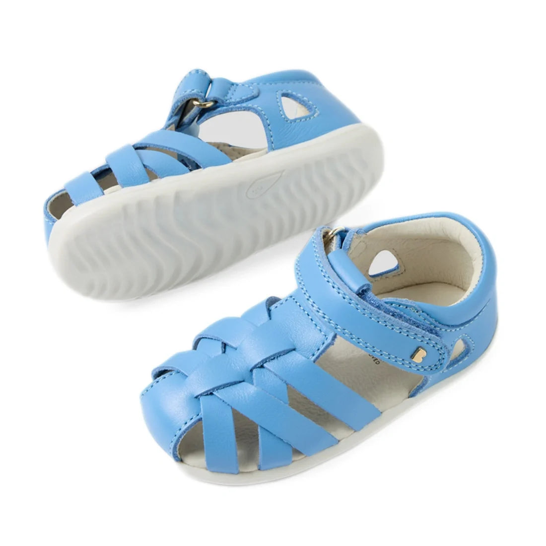 Two Baltic Sea Blue toddler leather Bobux Step Up SU Tropicana II Closed Toe Sandals with white soles. One sandal is positioned upright, revealing the top and side view, while the other is flipped to showcase the sole. The sandals have a woven design and 