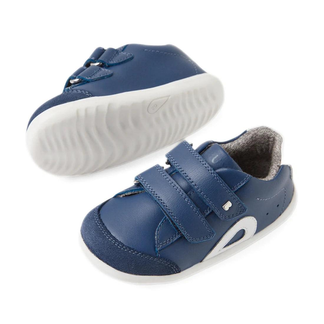 Bobux SU Step Up Blaze Leather Toddlers Sneaker in Vintage Indigo features quick-drying blue leather uppers, white flexible soles, easy Velcro straps, and a soft grey lining. Shown with one sneaker upright and the other on its side to display the bendable
