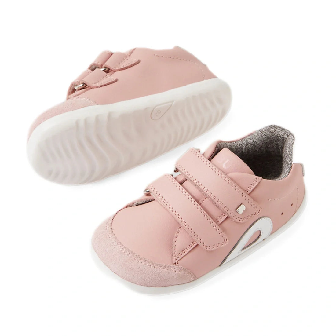 A pair of Bobux SU Step Up Blaze Leather Toddlers Sneaker in Ballet Pink, featuring white soles, Velcro straps, and a gray inner lining—perfect first walking shoes for your little one. One sneaker is upright while the other lies on its side to showcase th