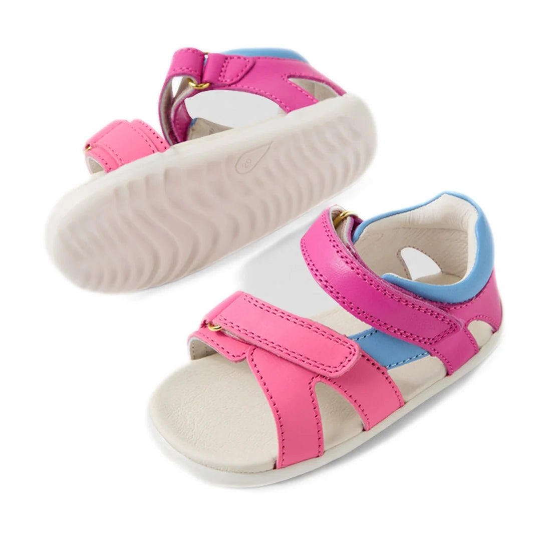 A pair of Bobux SU Step Up Dune First Walker Open Toe Sandals in Rose Bright Pink, featuring pink adjustable straps, blue accents, and a white sole. One sandal is upright while the other is shown from below to highlight the wavy texture of the sole. These