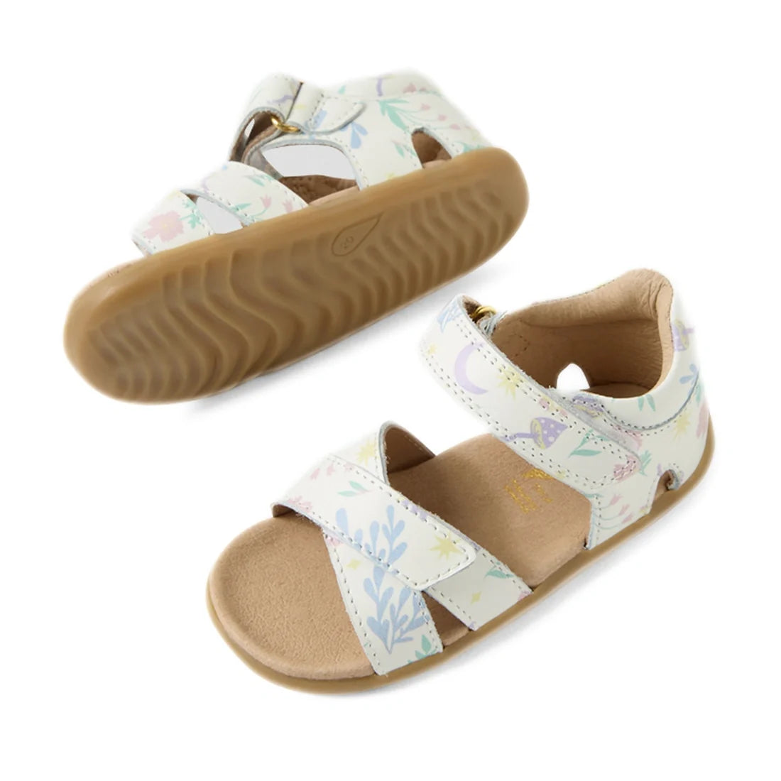 The Bobux Step Up Flora First Walker Sandal in White Floral features two children’s sandals with tan soles and white leather straps adorned with a pastel floral pattern. One sandal is upright, revealing the top and side, while the other is shown upside do